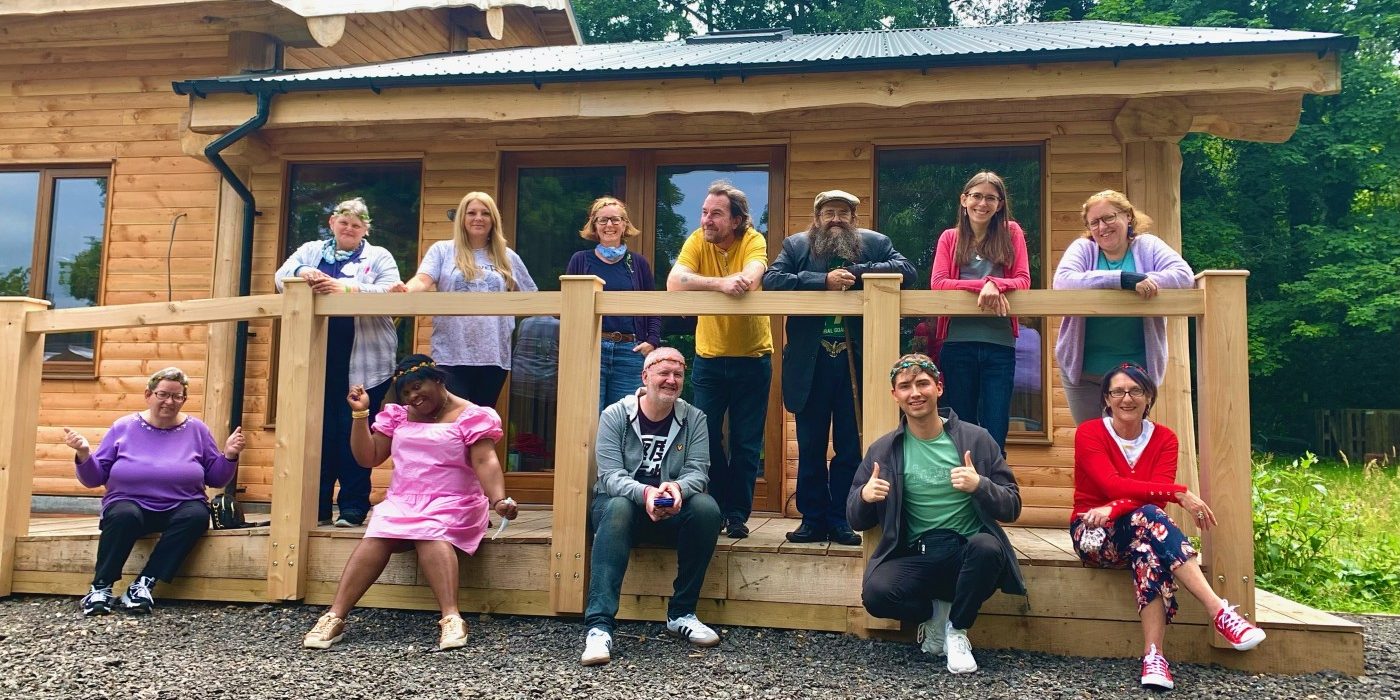 A group of 12 people, in two rows, outside a log cabin