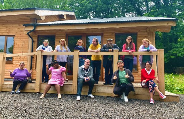 A group of 12 people, in two rows, outside a log cabin