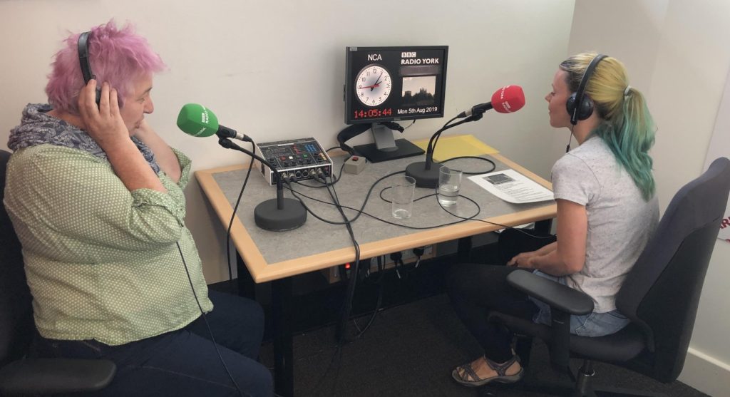 Mary Passeri & Sydnie Corley sit in a small radio studio, speaking into microphones.