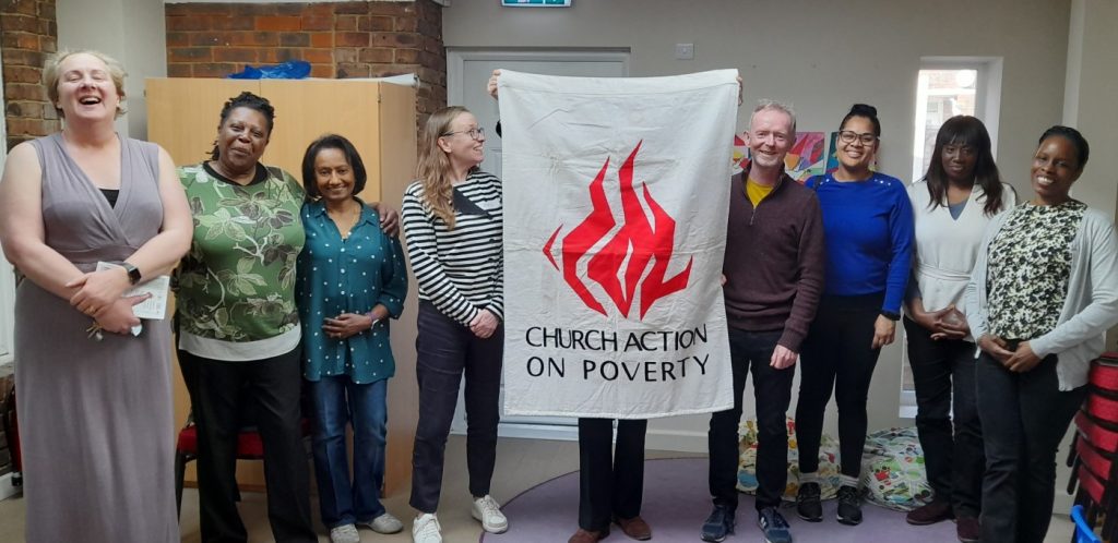 A group of 9 people in an office room, facing the camera,. The middle person is holding a vertical "Church Action on Poverty" banner.
