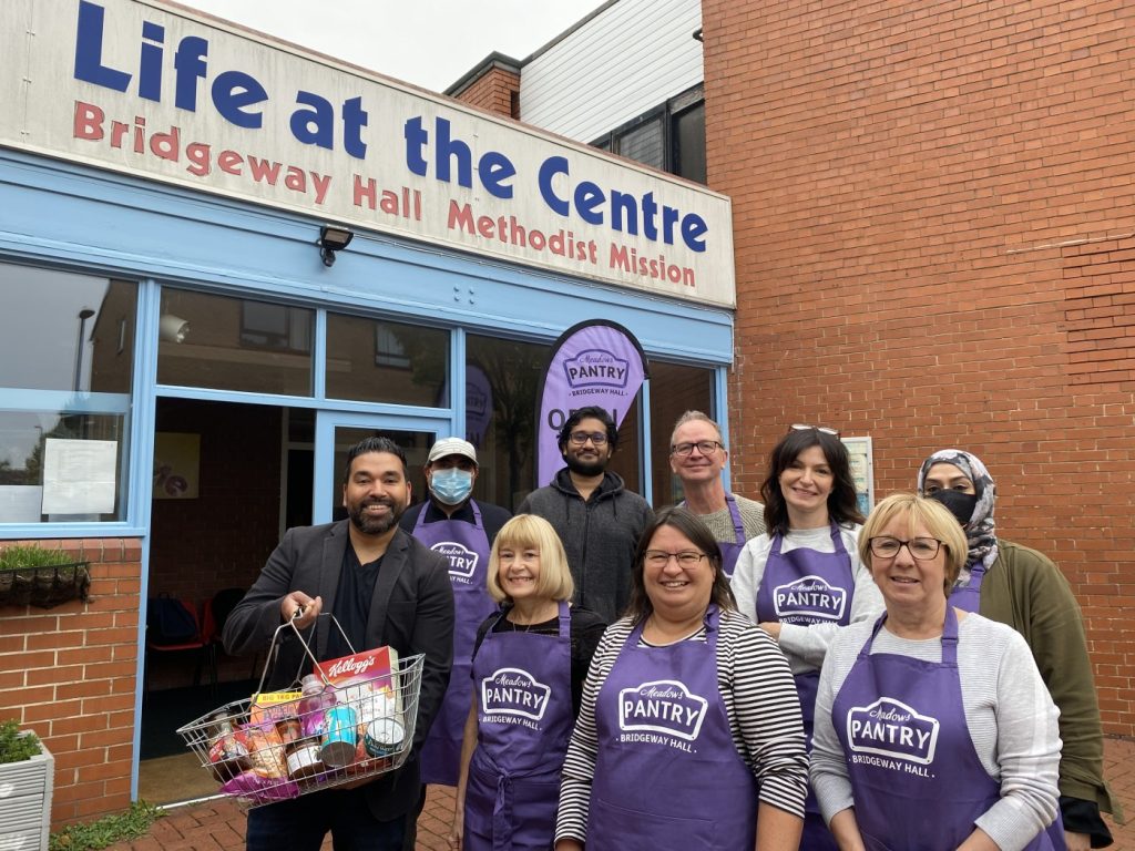 9 people outside a brick building, Nottingham's first Your Local Pantry. Most are in purple aprons; one is holding a basket of food.