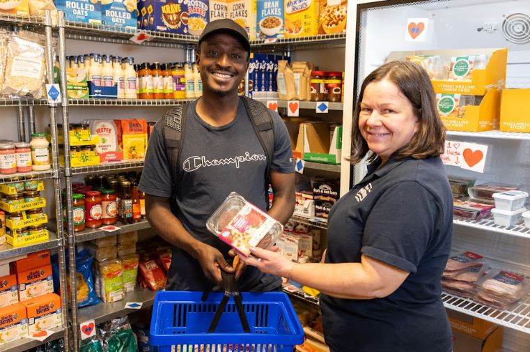 A Pantry member holds a basket beside a volunteer; both smile towards the camera