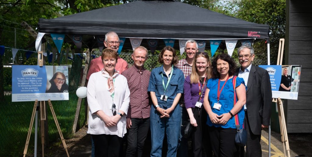 A posed line-up of 8 people in front of a gazebo and Your Local Pantry signs