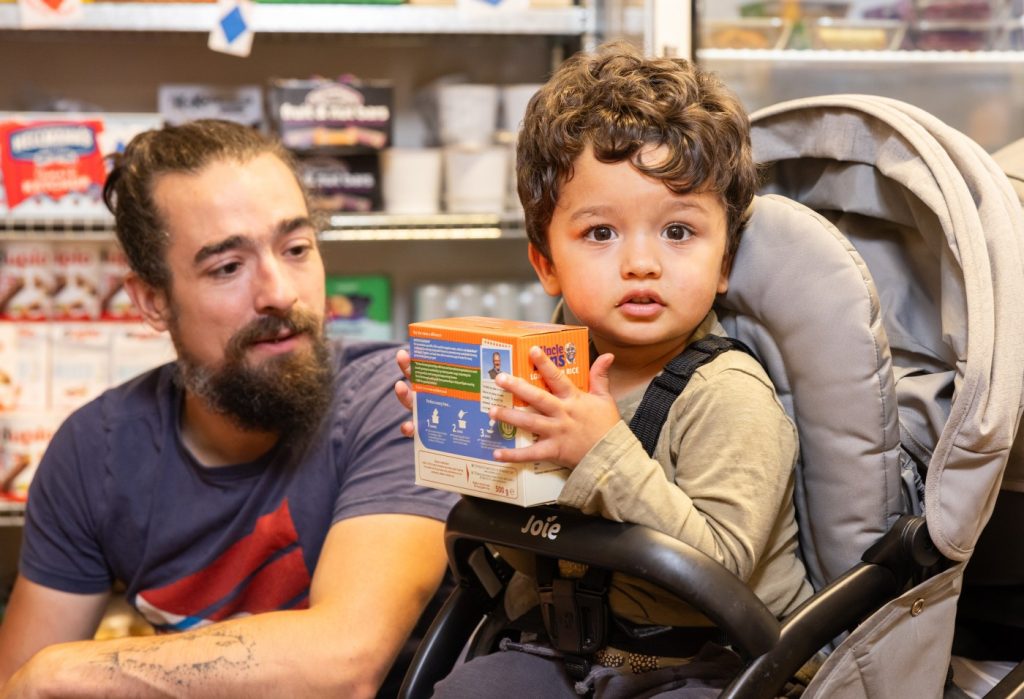 A toddler in a pushchair holds a box of rice, at a Your Local Pantry