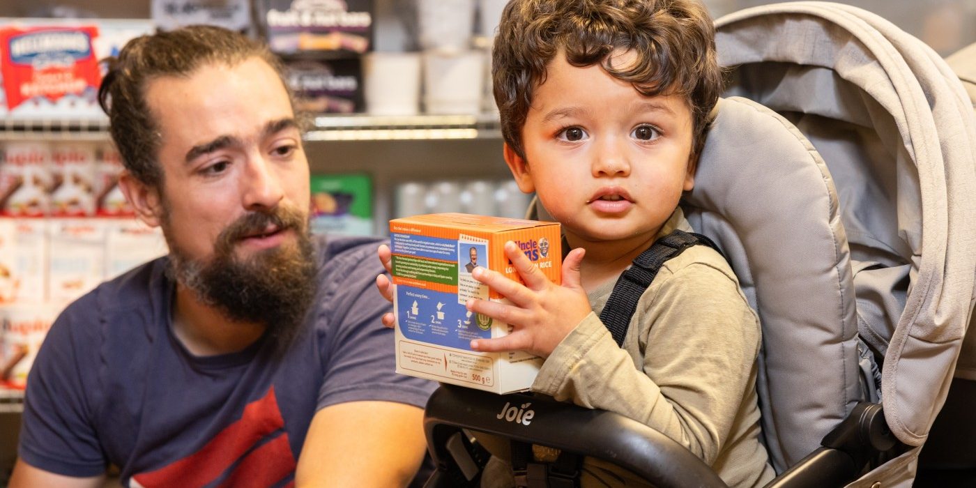 A toddler in a pushchair holds a box of rice, at a Your Local Pantry