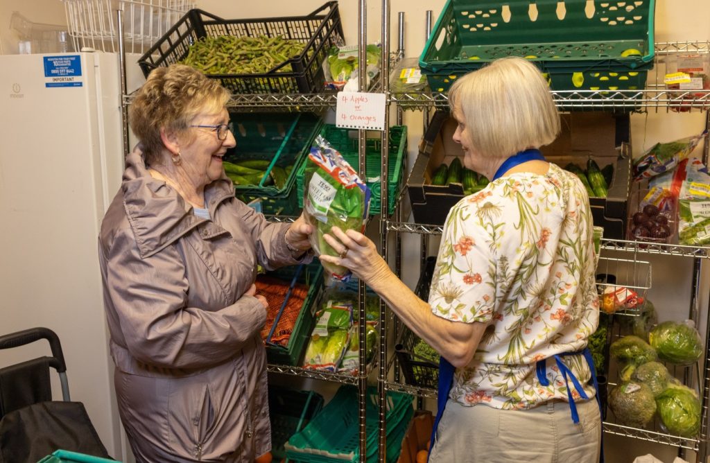 A woman takes a bag of salad from a shelf, while chatting to a volunteer.