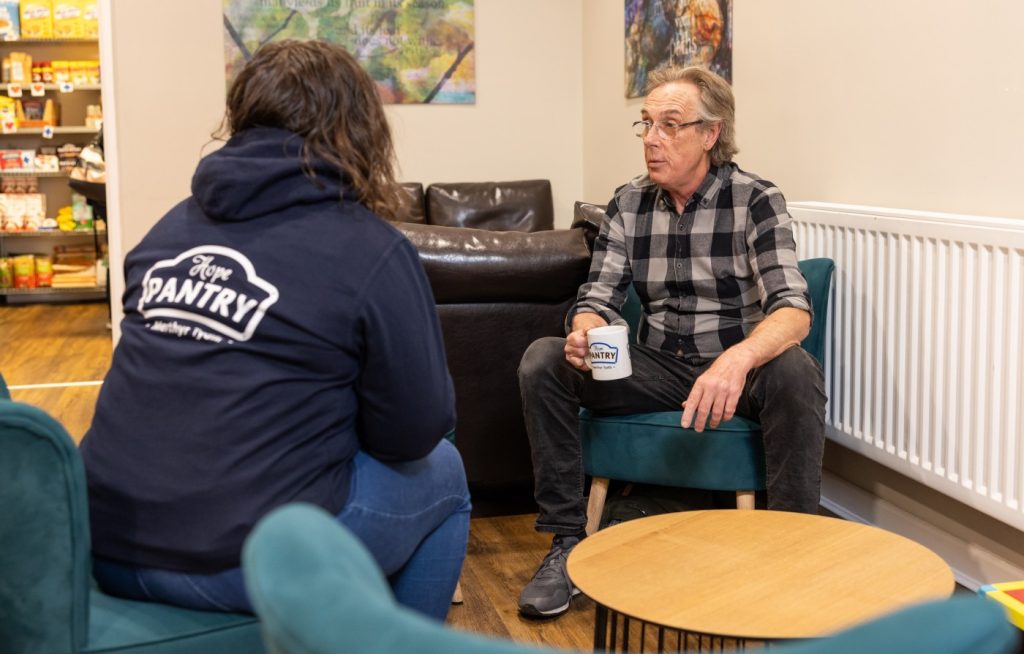 A volunteer in a Your Local Pantry hoody chats to a member. They are sitting beside a coffee table, the volunteer with her back to the camera, the member facing it.