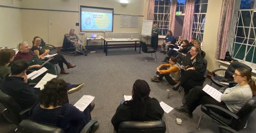 About a dozen people, sitting in a circle in a training room, with a projector screen at the front.