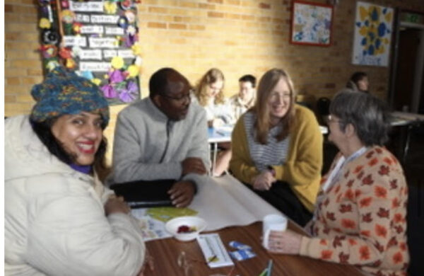 Four people at a table in a church hall. One is looking at the camera, the others are chatting. More people are further in the background. Sunshine is hitting the back wall.