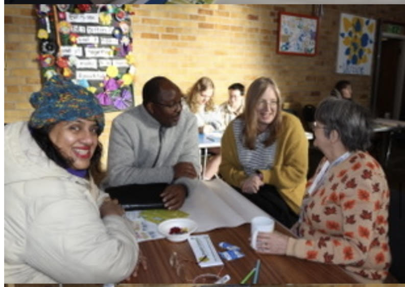 Four people at a table in a church hall. One is looking at the camera, the others are chatting. More people are further in the background. Sunshine is hitting the back wall.