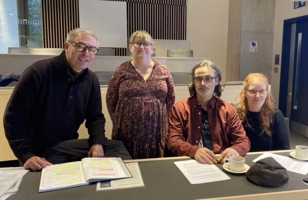Four people behind a desk, looking at the camera. Two are standing; two sitting.
