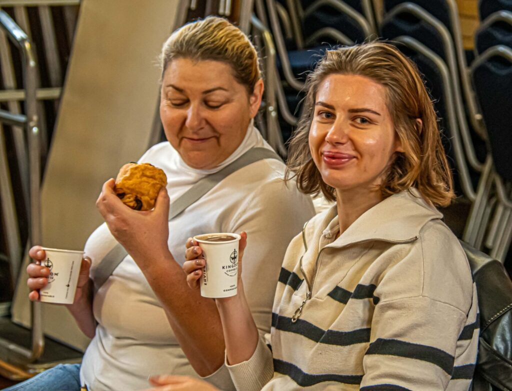 Two women sitting side by side, one with a hot drink and one with a pastry