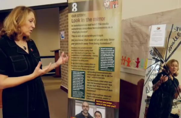A woman stands beside a banner about homelessness, facing a mirror with words written on it.