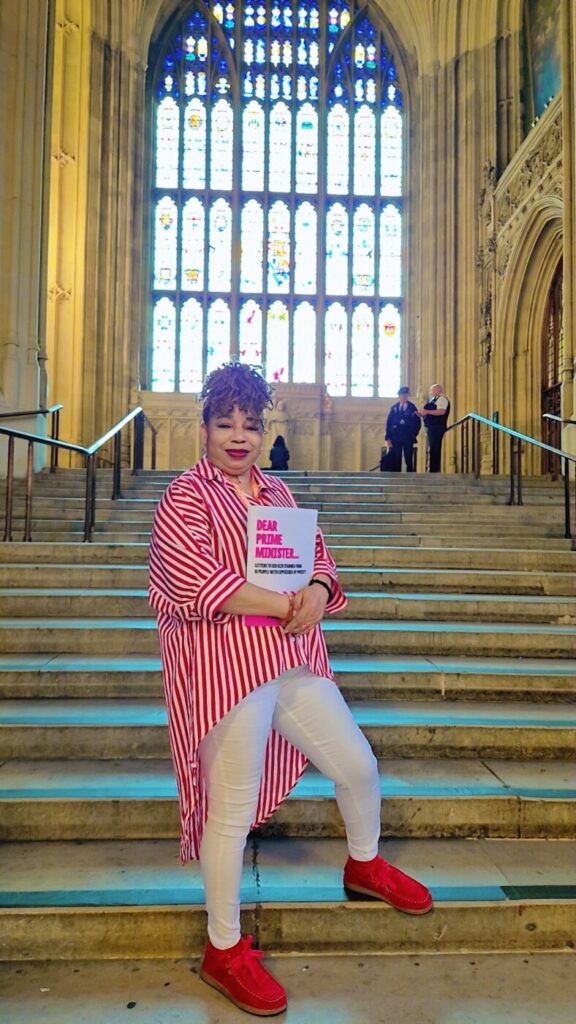 A portrait photo of Glory holding the "Dear Prime Minister" booklet, standing on a staircase in the Houses of Parliament.