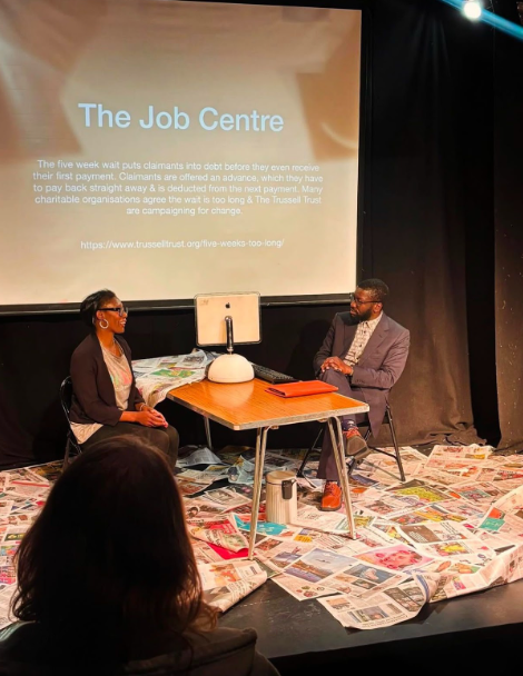 Two actors on a stage sit on opposite sides of a simple table. Newspaper pages are scattered across the floor. The backdrop reads: "The Job Centre"