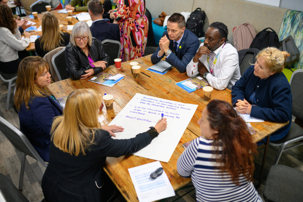 People sitting at tables, with one person writing on a large flip-chart sheet on the table