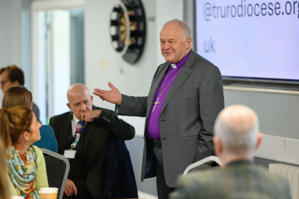 A clergyman in a purple shirt is standing speaking in a meeting room, with other people sitting at tables