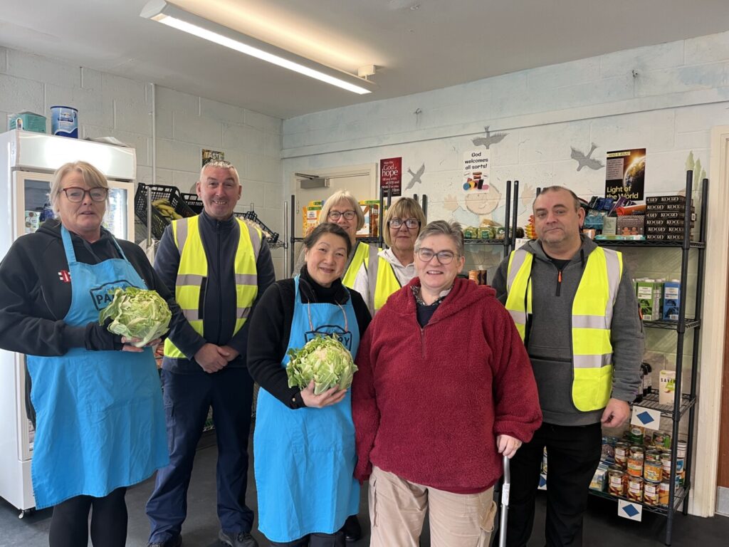 The team at Dingle Pantry. A woman in a red fleece stands with volunteers in blue aprons or high-vis jackets, in front of shelves of food.